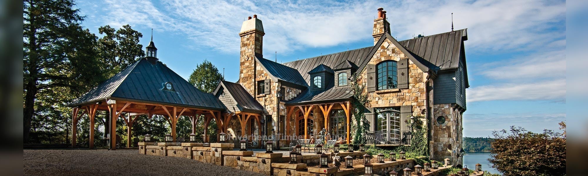 timber frame home with multi colored tan stone exterior with outdoor timber frame area with a lake in the background